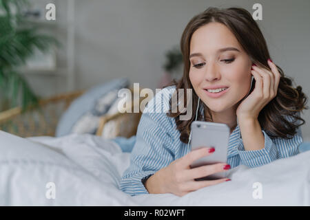 Bella donna ascolta musica, rende la chiamata video, risiede nel comodo letto in mattina, vestito casualmente, ha sorriso affascinante, pone in camera da letto. Persone, r Foto Stock