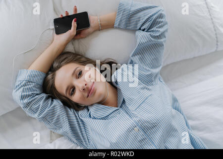 Concetto di rilassamento. Bella ragazza con felice espressione soddisfatta, vestito in pigiama, appoggia in letto, detiene smart phone, ascolta musica piacevole in earp Foto Stock
