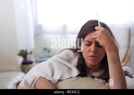 Giovani caucasian brunette ill donna con mal di testa e dolori di pancia, febbre, con le mani sulla testa e con coperta e termometro con atteggiamento di malattia, Foto Stock