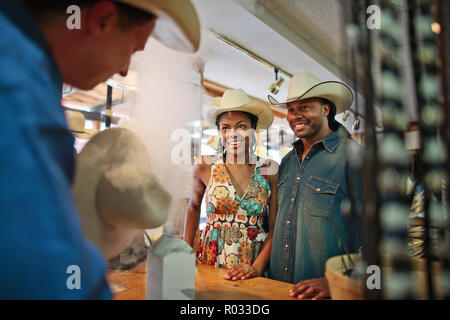 Sorridente coppia giovane guarda un artigiano mescolare la polvere come egli mette la rifinitura di un cappello da cowboy. Foto Stock