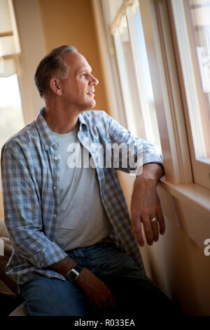 Uomo maturo guarda pensieroso fuori da una finestra piena di sole. Foto Stock