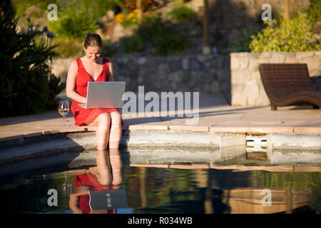 Metà donna adulta Dondolando i suoi piedi in piscina mentre su un laptop. Foto Stock