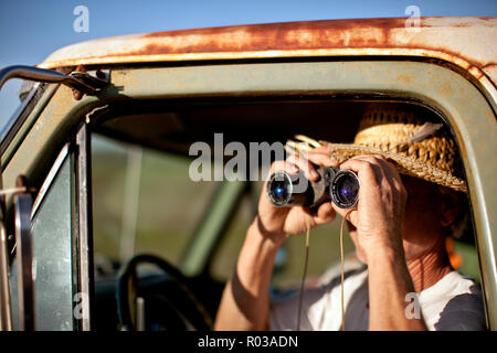 Uomo seduto in un pick-up truck guardando attraverso il binocolo. Foto Stock