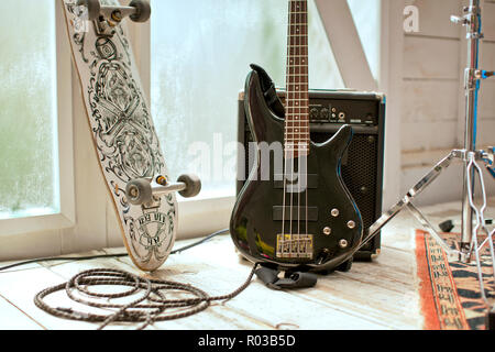 Skateboard accanto ad una chitarra basso e un amplificatore su un pavimento di legno duro. Foto Stock