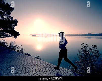 Tall slim è uomo che corre lungo il lago in posizione di parcheggio nella soleggiata lo spuntar del giorno. Misty del livello dell'acqua. Foto Stock