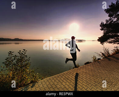 Tall slim è uomo che corre lungo il lago in posizione di parcheggio nella soleggiata lo spuntar del giorno. Misty del livello dell'acqua. Foto Stock