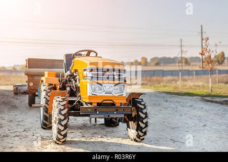 Mini trattore rosso con rimorchio in piedi vicino hangar edificio in azienda durante il tramonto o l'alba. Piccole macchine agricole. Paese rurale scena. Foto Stock