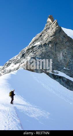 L'alpinista in piedi su una neve e ghiaccio ridge sotto una roccia taglienti ago picco di montagna sotto un cielo blu Foto Stock