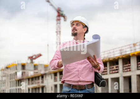 Ritratto di architetto al lavoro con il casco in un sito in costruzione, legge il piano, progetti di carta Foto Stock