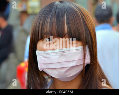 Giovane donna di Taiwan con bellissimi occhi orientali e fringe indossa un rosa chirurgico monouso lobo maschera facciale. Foto Stock