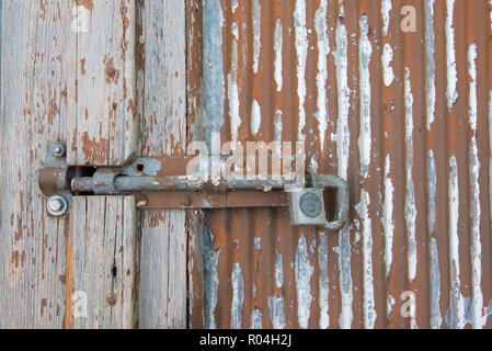 Un arrugginito dipinto bullone scorrevole è fissato con un lucchetto Lockwood su un vecchio in legno e acciaio zincato porta con peeling vernice rossa Foto Stock