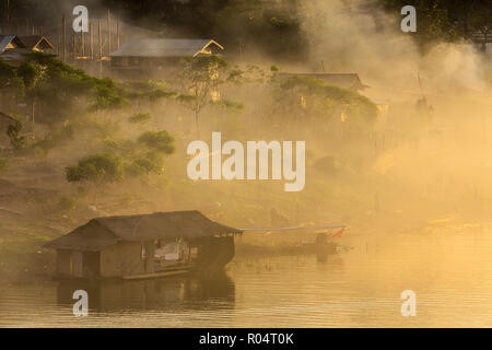 Misty tramonto tropicale sul lago con barca e casa di legno in Sangklaburi, Thailandia Foto Stock