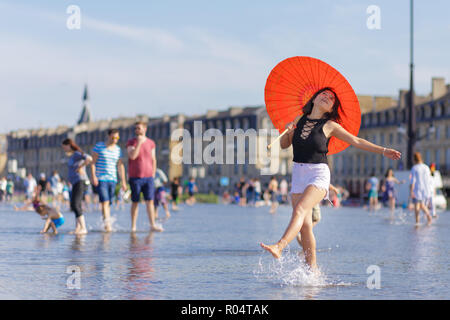 Donna asiatica con ombrello, ballare e spruzzi di specchio di acqua in place de la Bourse a Bordeaux, Francia Foto Stock