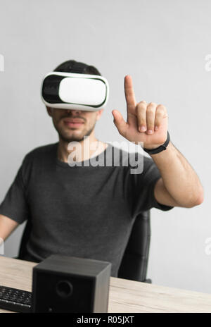 Giovane uomo che indossa la realtà virtuale gli occhiali di protezione auricolare e seduta in ufficio contro il computer. Il collegamento, la tecnologia di nuova generazione. Uomo cercando di toccare gli oggetti o il Controllo VR con una mano. Foto Stock