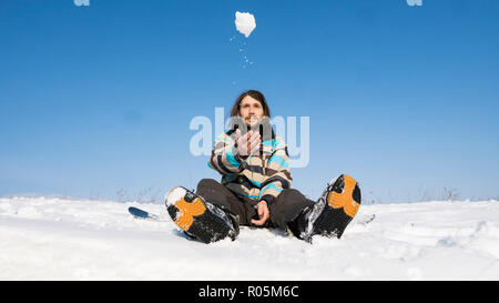 Snowboarder con capelli lunghi seduto su una di snowboard e di lanciare una palla di neve nell'aria. Extreme sport d'inverno. Foto Stock