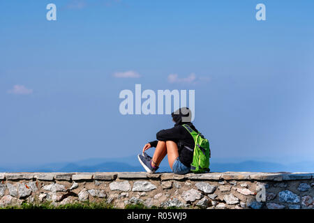 Un uomo con un zaino verde siede su un muro di pietra e rilassa. Foto Stock