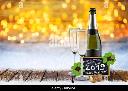 Bottiglia di champagne con bicchieri di sughero lavagna e felice anno nuovo 2019 biglietto di auguri su assi di legno di fronte oro brillante bokeh calda luce bac Foto Stock
