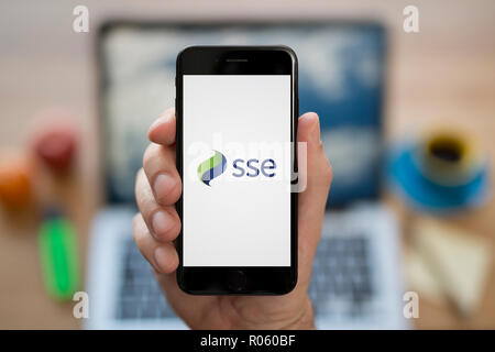 Un uomo guarda al suo iPhone che visualizza il logo di SSE, mentre se ne sta seduto alla sua scrivania per computer (solo uso editoriale). Foto Stock