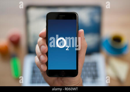 Un uomo guarda al suo iPhone che consente di visualizzare l'O2 logo, mentre se ne sta seduto alla sua scrivania per computer (solo uso editoriale). Foto Stock