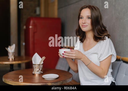 Piuttosto spensierato giovane donna in bianco informale di t-shirt, ha volto felice, bevande caffè aromatico, trascorre il tempo libero nel ristorante, siede al legno tondo tabl Foto Stock