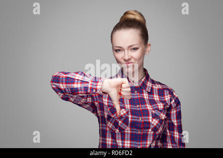 Infelice ragazza triste con rosa Maglietta a scacchi, raccolti bun acconciatura e trucco permanente e guardando la telecamera con faccia insoddisfatta e il pollice verso il basso. i Foto Stock