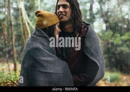 Giovane avvolto in una coperta sotto la pioggia all'esterno. Giovane uomo e donna avvolta in un plaid in piedi sotto la pioggia al parco. Foto Stock