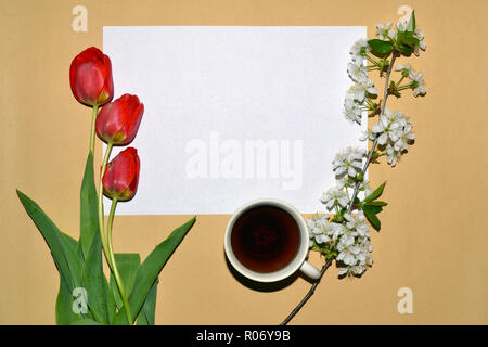 Una cornice di fiori bianchi e tulipani rossi intorno a un foglio di carta bianco e una tazza di tè. Foto Stock