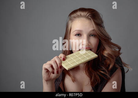 Giovane bella donna di mangiare il cioccolato bianco. Giovane biondo con trucco naturale divertendosi e mangiando cioccolato e sfondo grigio, copia dello spazio. Foto Stock