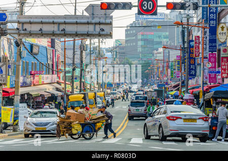 Un uomo spinge una mano carrello carico di cartone riciclato attraverso un passaggio pedonale attraverso una strada trafficata in Busan, Corea del Sud. Foto Stock