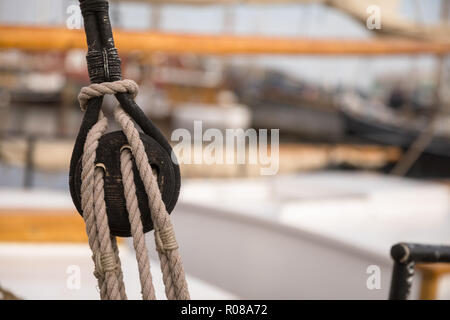 Puleggia per vele e corde di legno su una vecchia barca vela, con vela e altre barche nel porto, morbido e fuori fuoco in background. Foto Stock