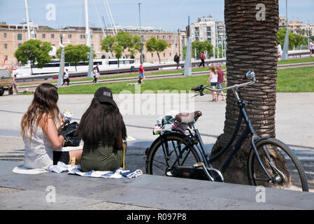 Due giovani donne seduta sul marciapiede e a pranzo con una bicicletta dai loro lati, Barcellona, Spagna Foto Stock