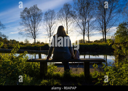 Donna seduta su una panchina di una tranquil riverside scena con un cielo blu riflessa nell'acqua e alberi e lussureggianti cespugli di rivestimento del lungofiume. Foto Stock