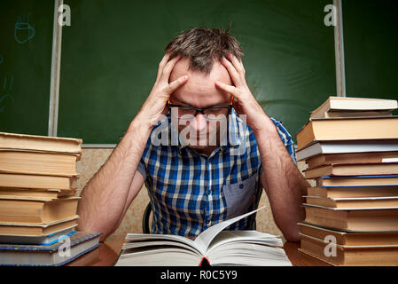 Un disheveled stanco barba lunga giovane uomo con gli occhiali tiene la sua testa e legge un libro a tavola con pile di libri contro lo sfondo di un blackb Foto Stock