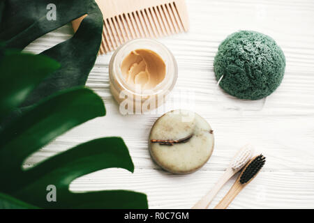 Eco Friendly di bamboo naturale spazzolini da denti, shampoo bar, dentifricio in vetro, spazzola in legno e spugna konjaku su legno bianco verde con foglie di monstera. Foto Stock