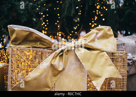 Golden confezioni regalo sotto l'albero di Natale in città europea centro. Big presenta con archetti su sfondo di luci di Natale. Decorazioni di festa di strada di città Foto Stock