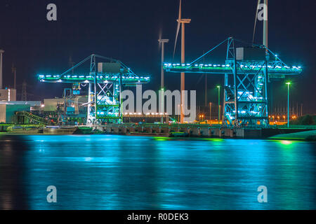 Wharf durante il tramonto con enormi gru moderne sotto illuminazione colorata e delle turbine a vento nel contesto Foto Stock