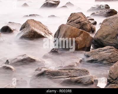 Esposizione lenta sfuma le onde nel mare che si rompono sulle rocce al tramonto Foto Stock