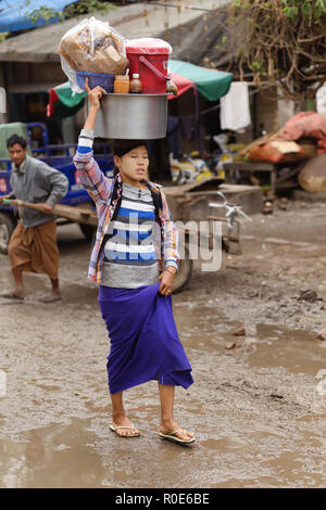 AMARAPURA, Myanmar, gennaio 17, 2015 : Una donna sta portando un grande contenitore metallico sulla sua testa, camminando nella strada sporca del mercato Zegyo, in Foto Stock