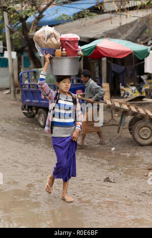 AMARAPURA, Myanmar, 17 gennaio 2015: è una donna che porta un sacco sul suo capo, camminando nella strada sporca del mercato Zegyo, a Mandalay, Myanmar (Bu Foto Stock