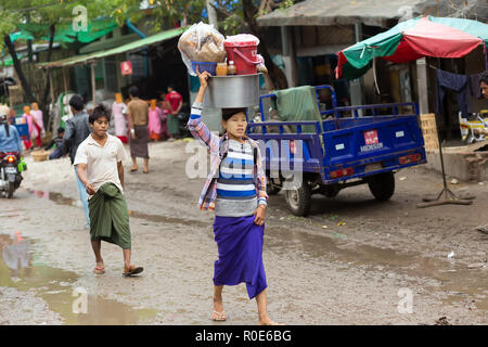 AMARAPURA, Myanmar, gennaio 17, 2015 : Una donna sta portando un grande contenitore metallico sulla sua testa, camminando nella strada sporca del mercato Zegyo, in Foto Stock