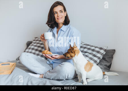 Ritratto di sognante brunette donna siede gambe incrociate su un comodo letto in camera da letto, guarda pensieroso a parte, detiene tazza di caffè e un croissant e lui Foto Stock