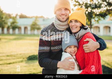 Posizione orizzontale Ritratto di giovane uomo bello nel cappello giallo e maglione caldo, abbracciare la moglie e la figlia, pongono alla telecamera mentre stand nel parco. T Foto Stock