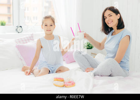 Ritratto di felice giovane madre e la sua piccola figlia di tenere bicchieri di latte shake, avere la colazione a letto, guardare positivamente al fotocamera, gode di weekend a Foto Stock