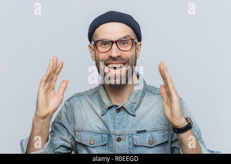 Bel uomo felice alza le mani con garantite, ha detto contentissimo espressione, guarda con piacere il viso, è folta barba e baffi. Felice l'uomo in eyewear Foto Stock