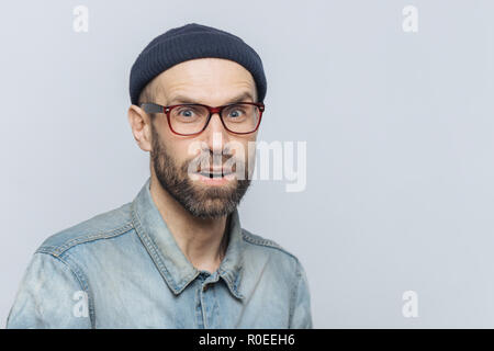 Elegante di mezza età con maschio sorpresi e curiosi espressione indossa occhiali, camicia di denim e hat, pone contro uno sfondo grigio con spazio copia fo Foto Stock