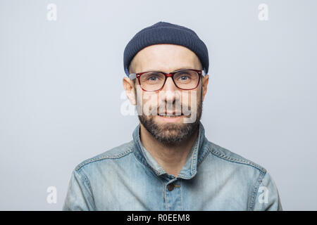 Ritratto di bello ed elegante giornalista maschio indossa occhiali, Elegante cappello e camicia denim, pone contro il bianco di sfondo per studio. Soddisfatto l uomo wit Foto Stock