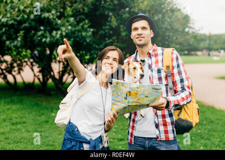 Bella donna con i capelli scuri che mostra per l uomo che ha perso in città. Bel ragazzo con cane avente viaggio nel parco, azienda mappa in mani chiedendo passerb Foto Stock