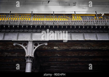 Servizio in giallo il treno su un vecchio ponte in acciaio con pilastro, in un giorno nuvoloso, con catenaria e un uccello nero Foto Stock