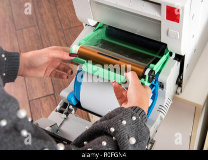 Donna cambiando il nastro su una stampante fotografica al di sopra di un nuovo rotolo di carta di pellicola in una vista ravvicinata della macchina e delle sue mani Foto Stock