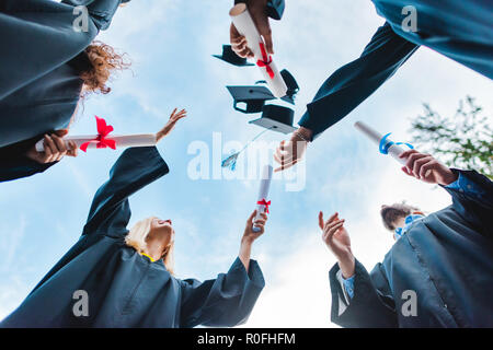 Vista dal basso del felice laureati multiculturale con diplomi di gettare i tappi con cielo blu su sfondo Foto Stock